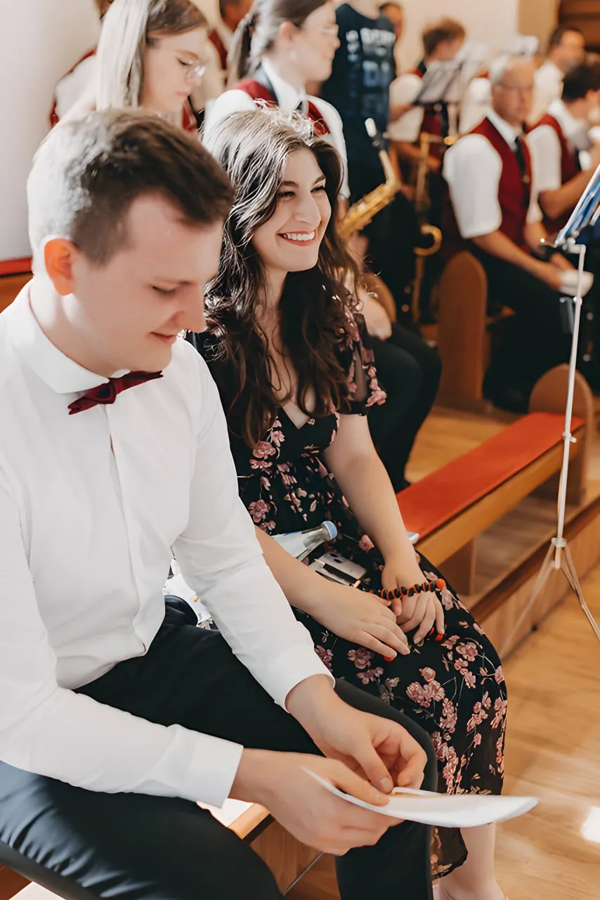 Dominik und Sophia sitzen lachend in einer Kirchenbank, im Hintergrund eine Blaskapelle